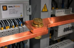A professional macro photograph capturing the interior of an industrial low-voltage electrical panel. In sharp focus, two bright orange-red copper busbars (شمش مس) are meticulously bolted together using a golden brass hexagonal head bolt, complete with a matching brass flat and spring washer. The background is softly blurred, showing technical electrical wiring, neatly arranged terminal blocks, and a clean corporate graphite-grey control board with subtle safety indicators. A warm, precise studio light gleams off the metallic brass and copper surfaces, creating a high-contrast engineering aesthetic. No sparks, highly realistic, 8k resolution, authentic B2B industrial environment